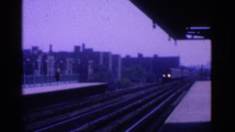 Rail tracks run beside a station platform toward a distant train, with urban buildings silhouetted in the purple-tinted 8mm frame.