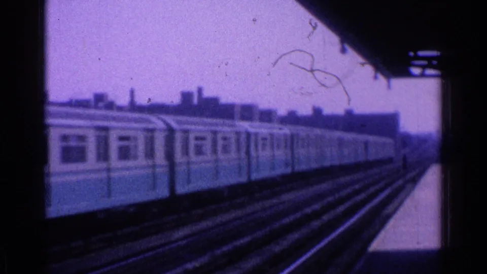 1965 Ohio 8mm frame showing the side of a light-colored train passing along elevated tracks with skyline behind it.
