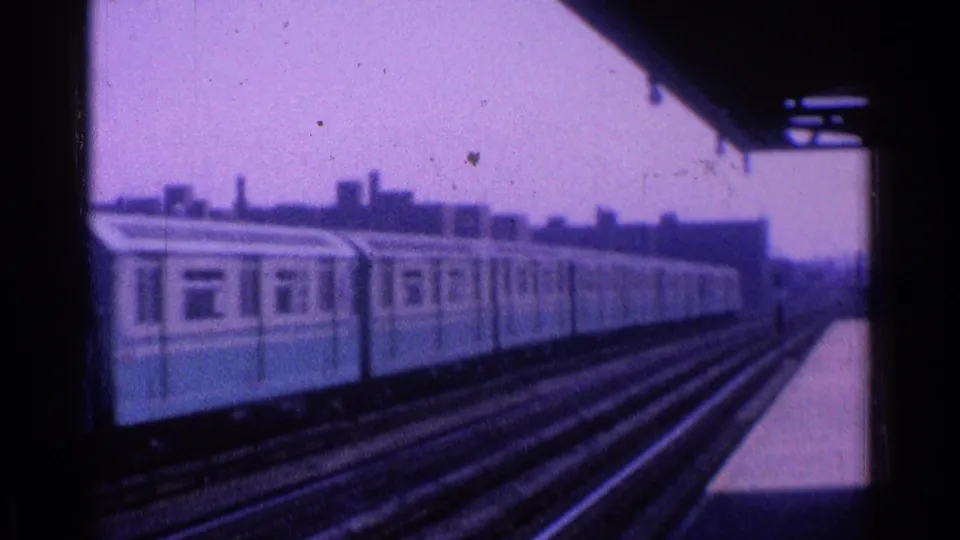 1965 Ohio home-movie frame of a train moving past an elevated station, with tracks and hazy skyline visible.