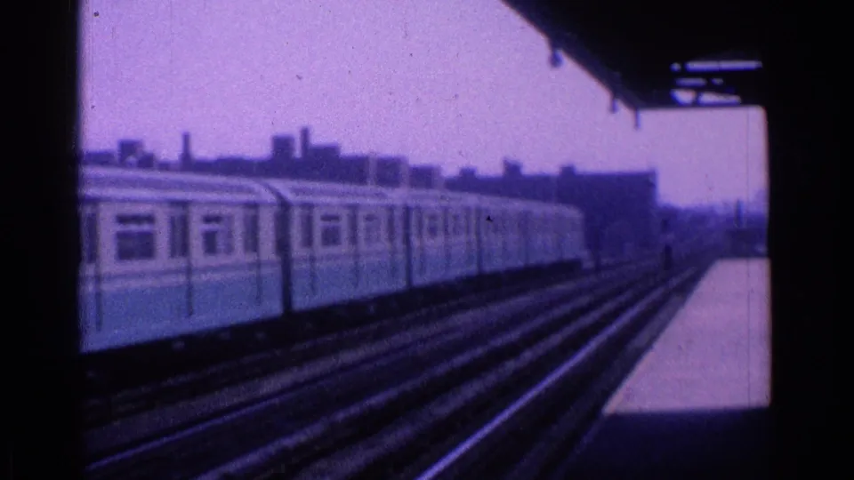 1965 Ohio home-movie frame showing a blue-and-cream passenger train along elevated city tracks beneath a station canopy.