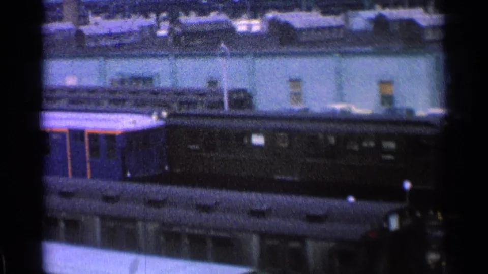 1965 Ohio 8mm frame showing a dark train car below station-side buildings and rooftop equipment, seen from a moving train.