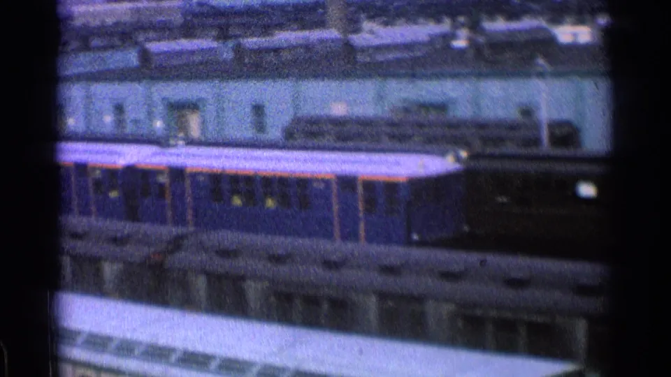1965 Ohio rail-yard view with a dark train car, station-side building, rooftop fixtures, and trackside structures in soft 8mm color.