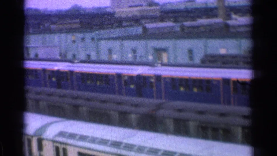1965 Ohio 8mm frame showing blue passenger cars, pale yard buildings, tracks, and dark film-frame edges in a rail yard.