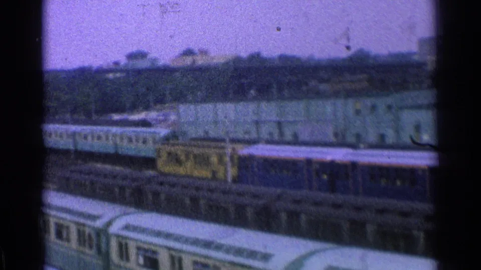 1965 Ohio home-movie frame with blue rail cars, a yellow car, pale green rolling stock, tracks, and industrial buildings.
