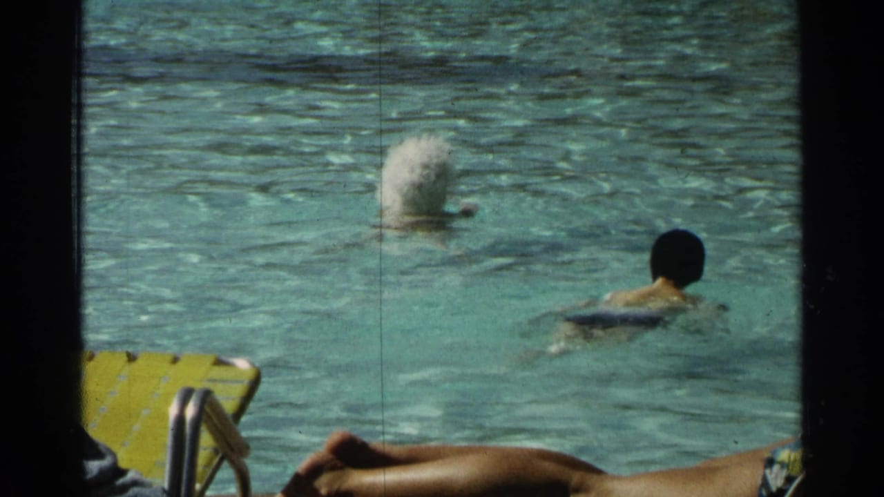 Swimmer in a turquoise resort pool near a splash and yellow lounge chairs.