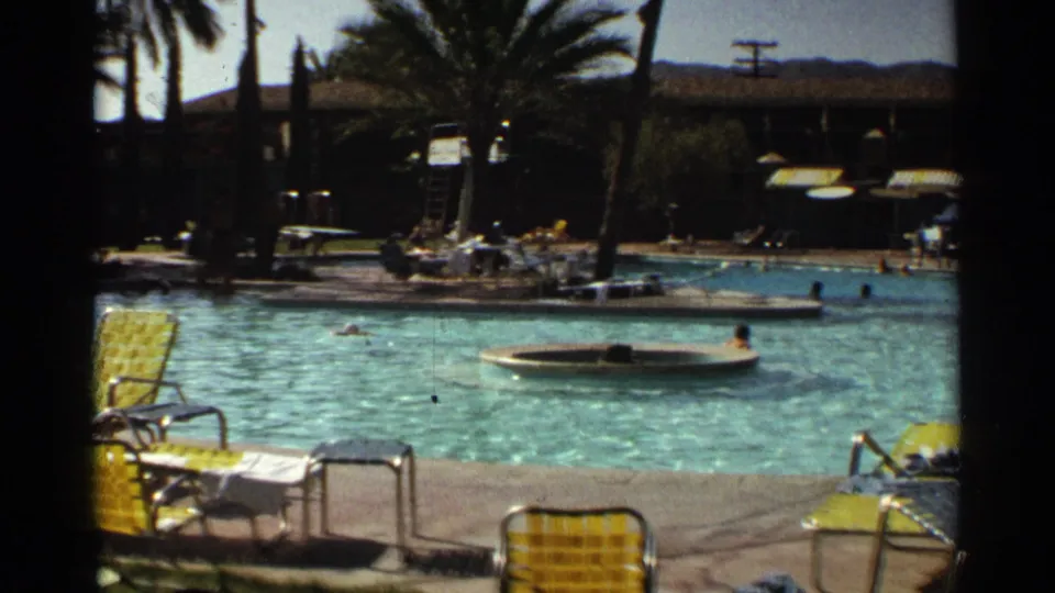 1968 Scottsdale resort pool with swimmers, yellow lounge chairs, palm trees, shaded buildings, and desert hills beyond.