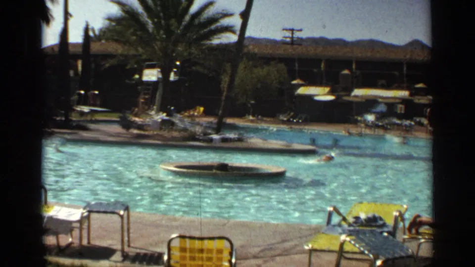 1968 Scottsdale poolside resort view with swimmers, yellow lounge chairs, palms, shaded structures, and distant hills.