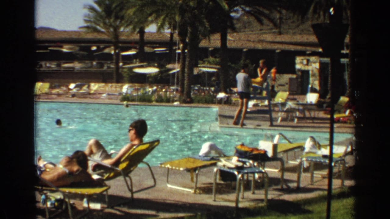 Poolside guests sit and stand near lounge chairs while swimmers move through the water.