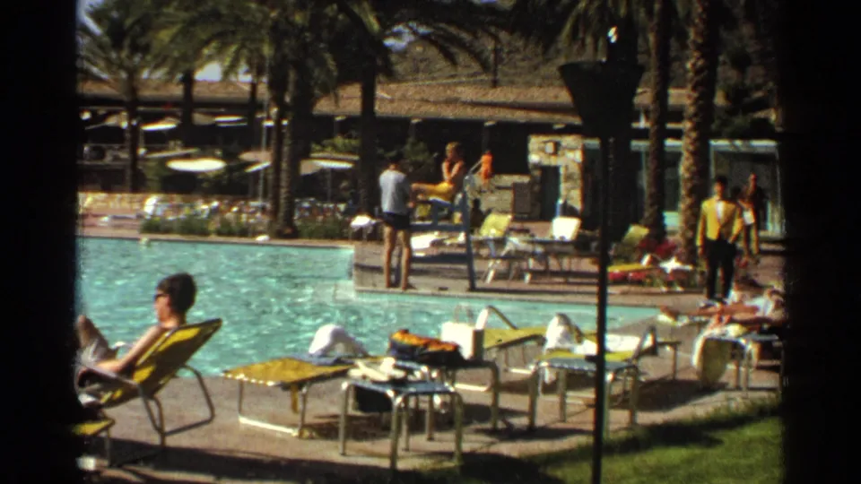 Scottsdale poolside home-movie frame from 1968 showing sunbathers, swimmers, yellow chairs, palm trees, and shaded resort buildings.