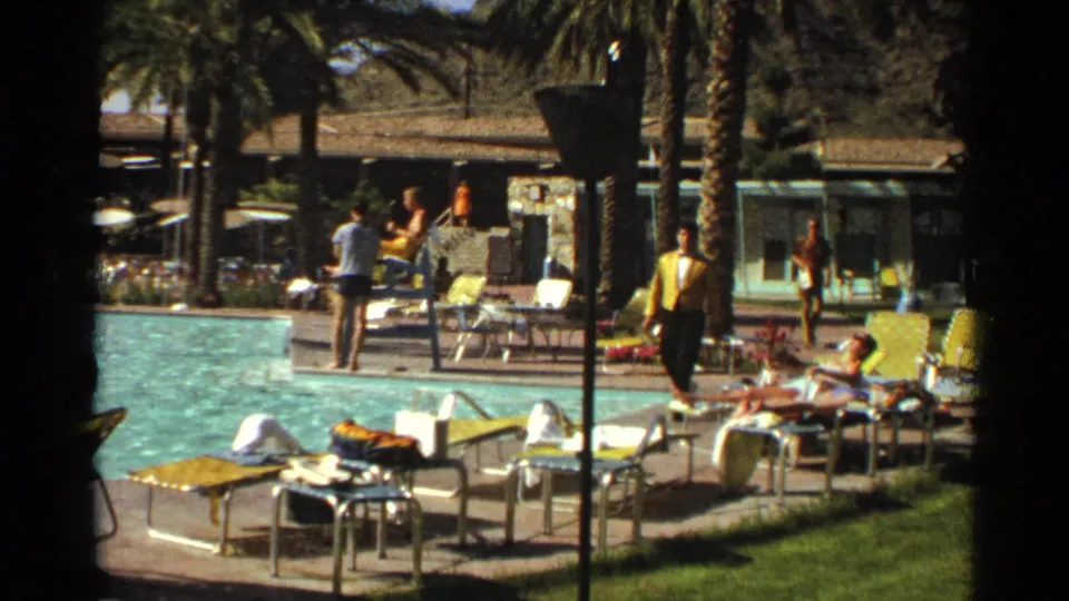 1968 Scottsdale resort pool deck with adults on yellow loungers, a person walking by palm trees, swimmers, and low buildings in the background.