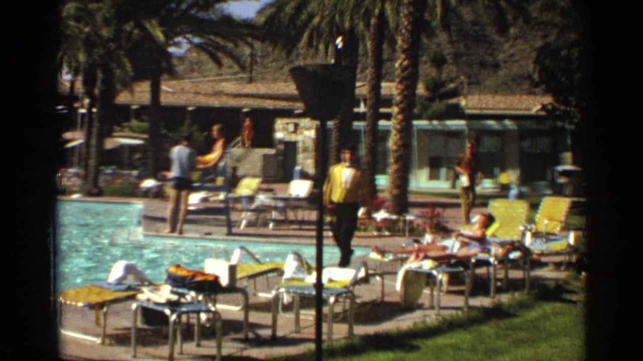 Poolside resort scene with lounge chairs, palm trees, guests, and a man in yellow walking near the water.