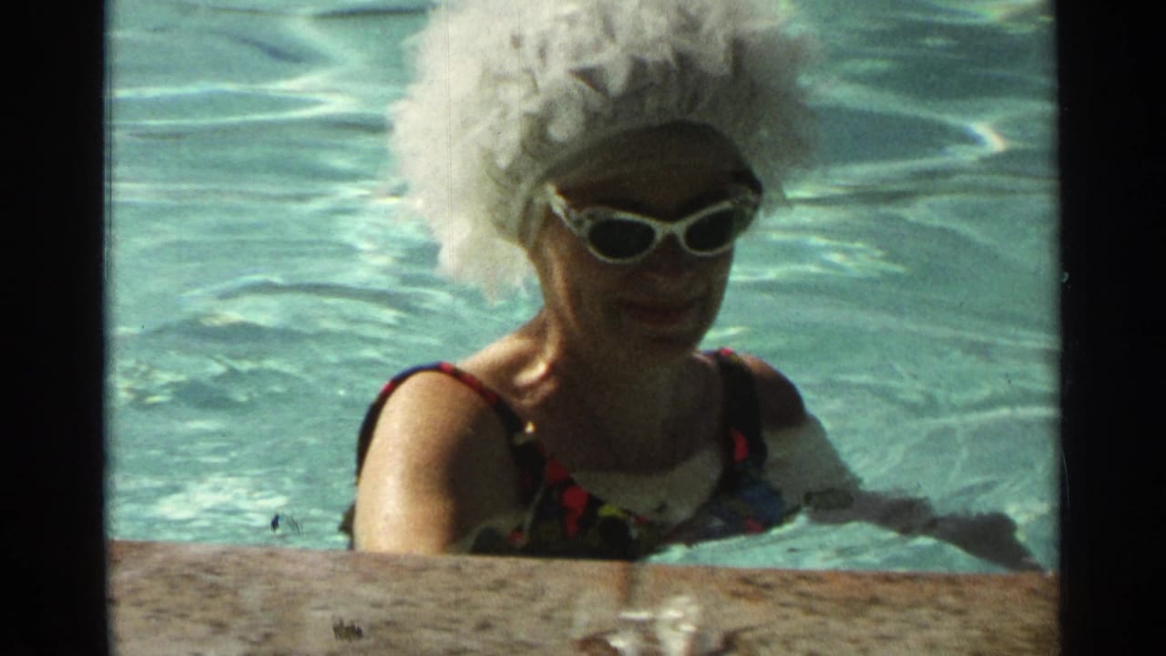 In Scottsdale, Arizona in 1968, a woman in a patterned swimsuit, white wig, and white sunglasses smiles in a blue swimming pool.