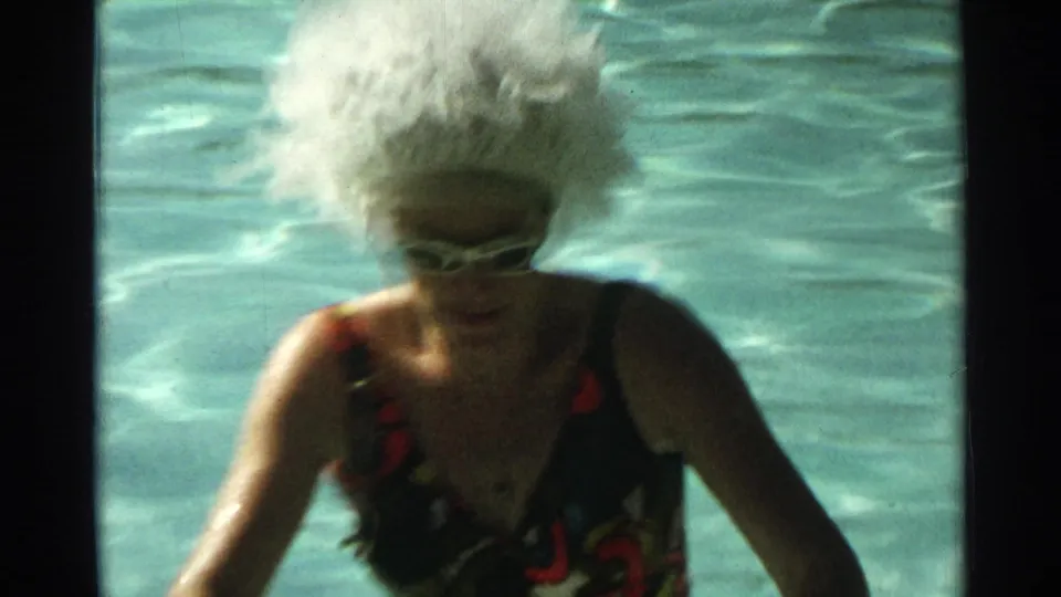 The woman leans forward in the Scottsdale pool, her white wig and dark patterned swimsuit visible in soft 8mm focus.