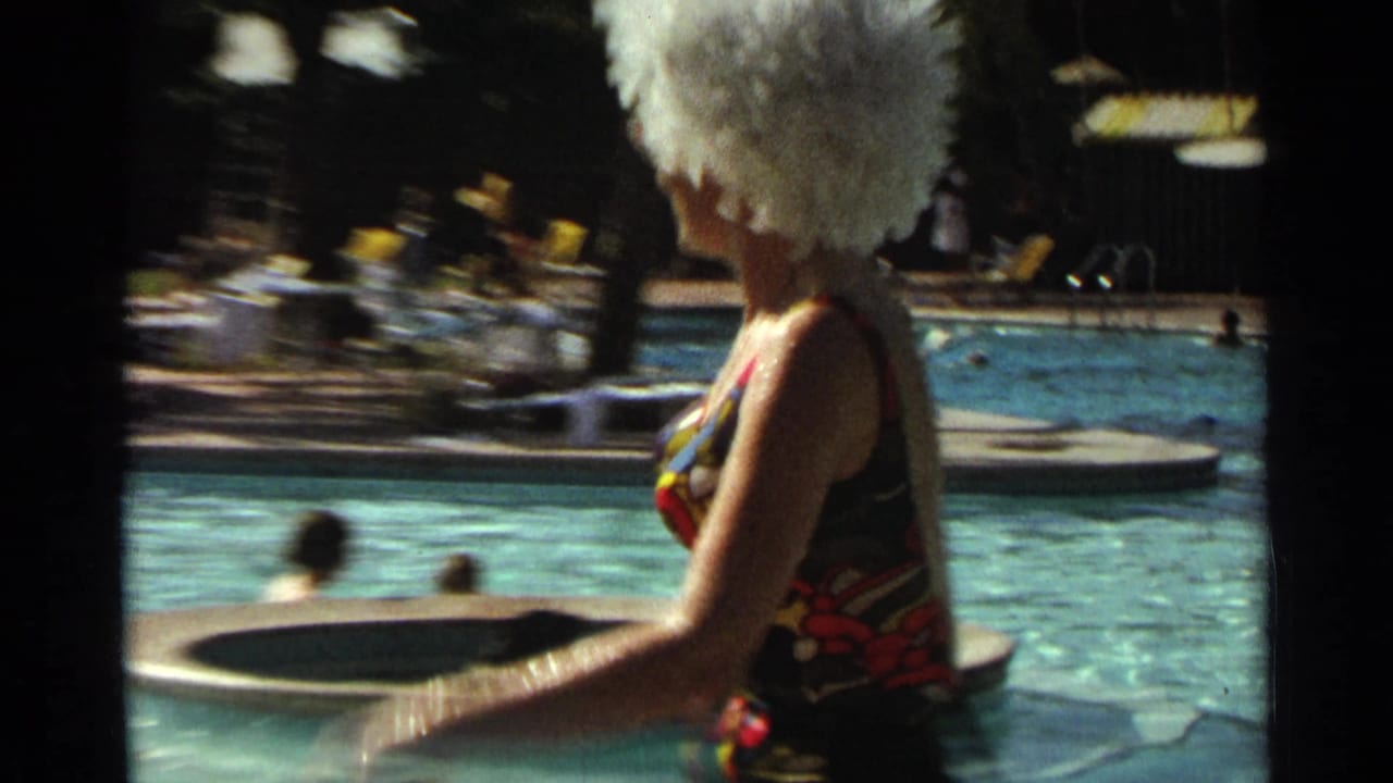 Woman in a colorful swimsuit and white wig walking beside a swimming pool.