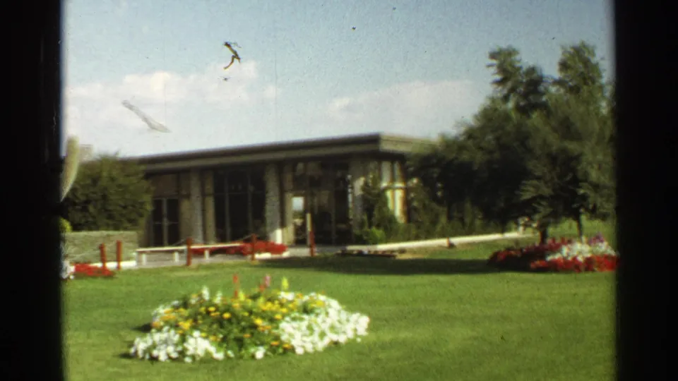 Scottsdale, Arizona frame of a low modernist building seen beyond lawn, flower beds, and red posts in 1968 footage.