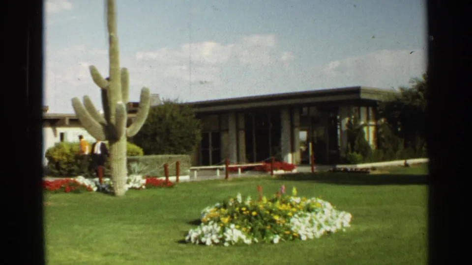 1968 Scottsdale frame with a prominent saguaro cactus, low building, flower beds, and two distant people on the lawn.