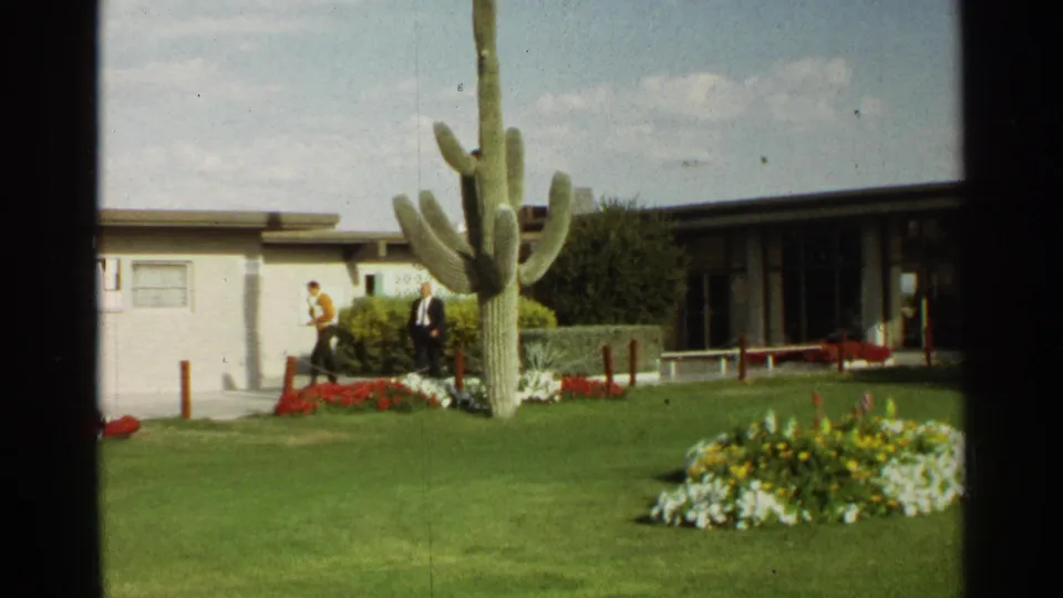 Scottsdale, Arizona home-movie frame showing two adults walking near a saguaro cactus beside a low building in 1968.