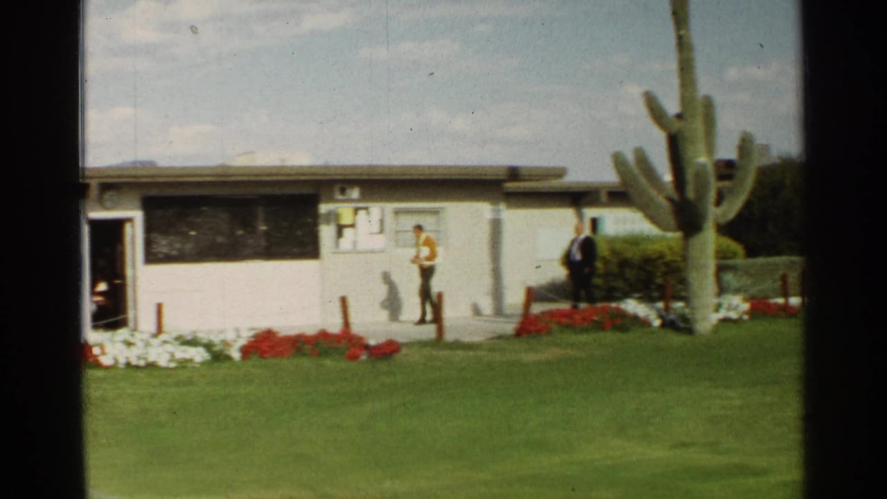 1968 Scottsdale frame of two men walking beside a low building, with desert landscaping and a large saguaro in the foreground.