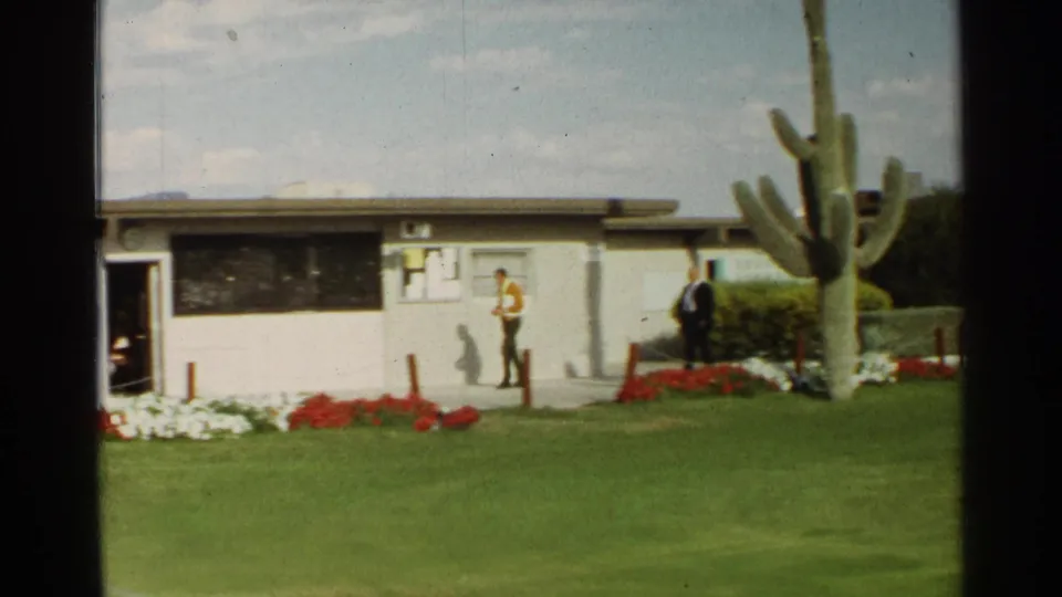 1968 Scottsdale frame of two men walking beside a low building, with desert landscaping and a large saguaro in the foreground.
