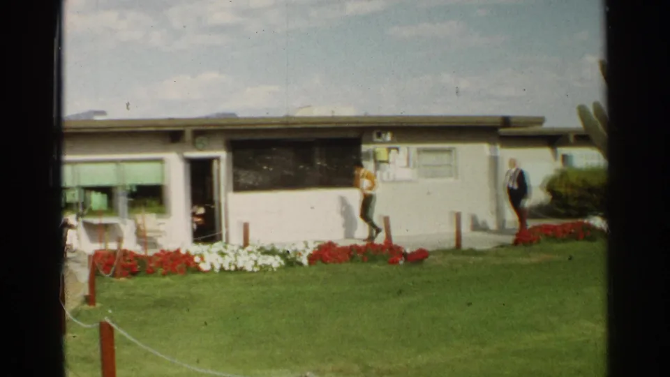 1968 Scottsdale home-movie frame with a man in a yellow shirt walking by a low building and another man near red flowers.