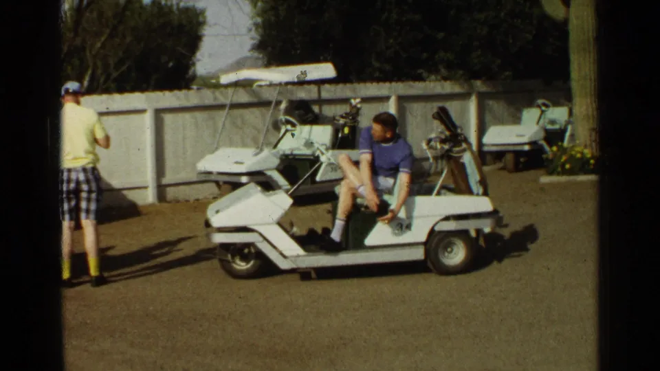 1968 Scottsdale frame showing a man seated in a white golf cart and another man standing nearby beside a fence and recreational equipment.
