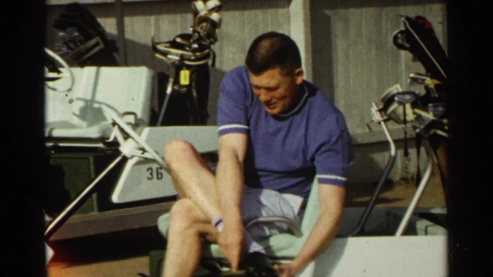 A seated golfer in a blue shirt adjusts his shoe on a white cart, with clubs and cart equipment behind him.