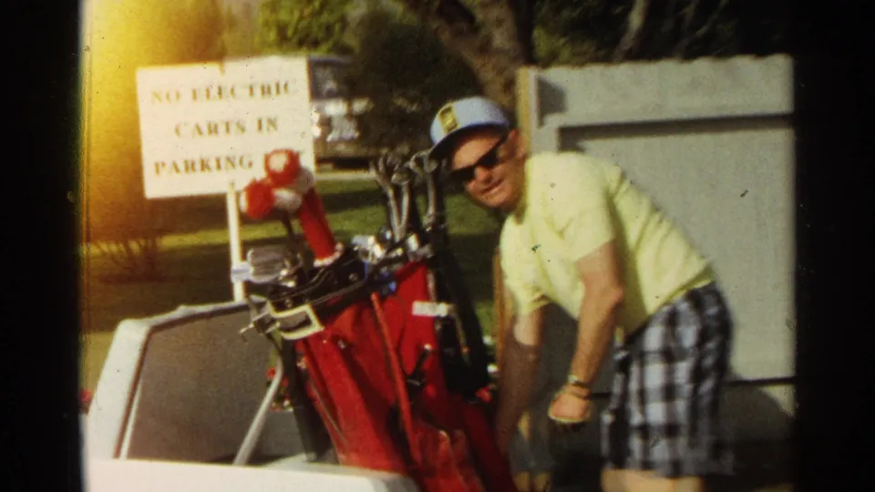 A man in a yellow shirt, plaid shorts, hat, and sunglasses stands near a red golf bag and a sign about electric carts.
