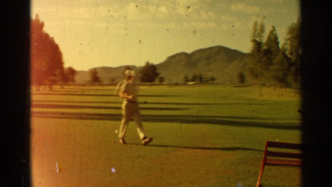 Golfer in a light outfit walking across a green with mountains and trees behind him.
