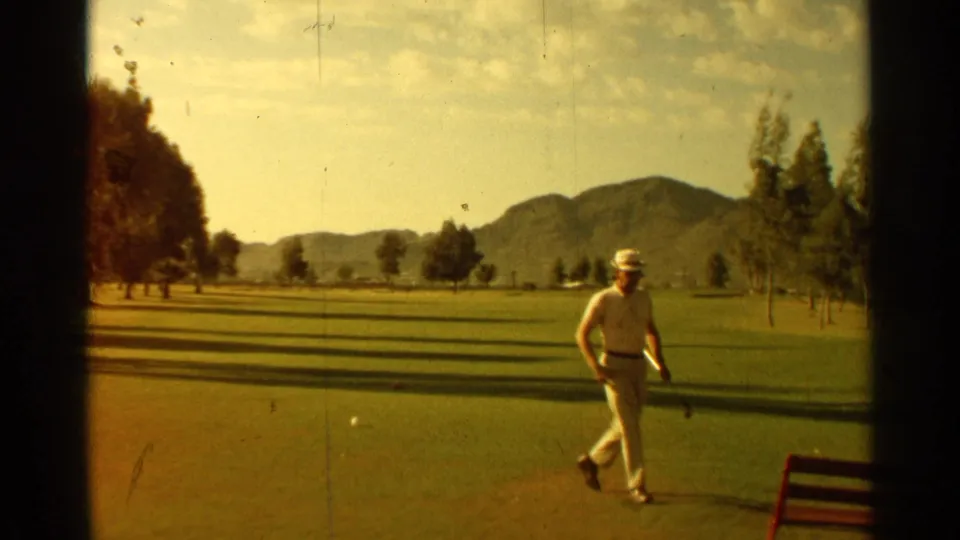 1968 Scottsdale home-movie frame of a golfer crossing a green, with open fairway, trees, mountains, and a red object at frame edge.