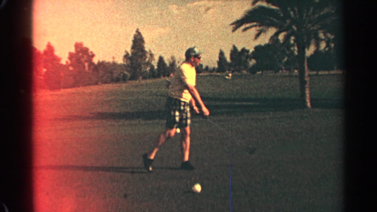 Man in light shirt and plaid shorts approaches a golf ball on a fairway.