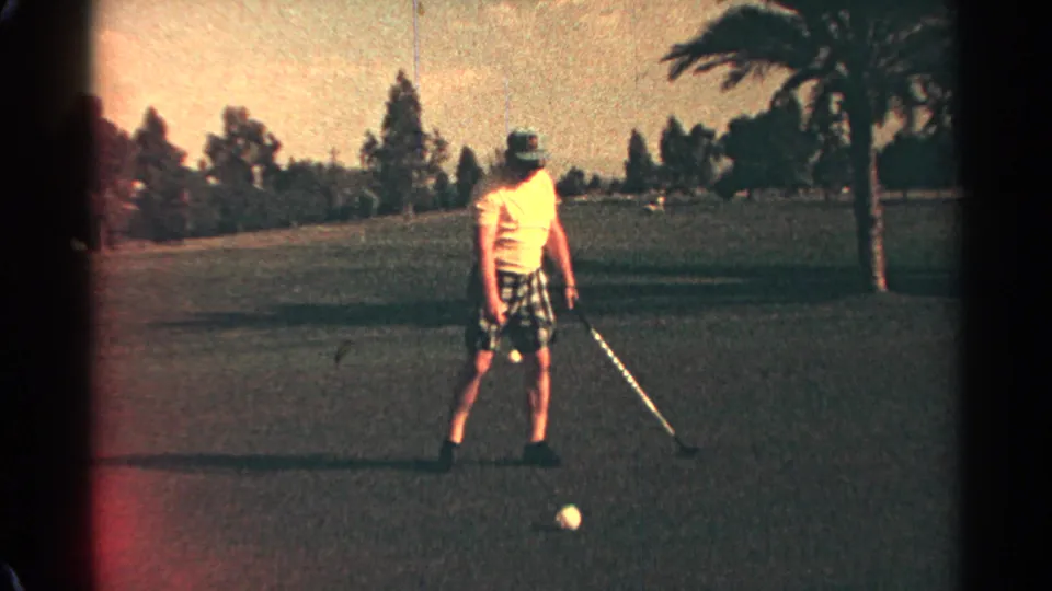A male golfer pauses near a white ball on a Scottsdale course, with palm and tree-lined fairway visible in faded 8mm color.