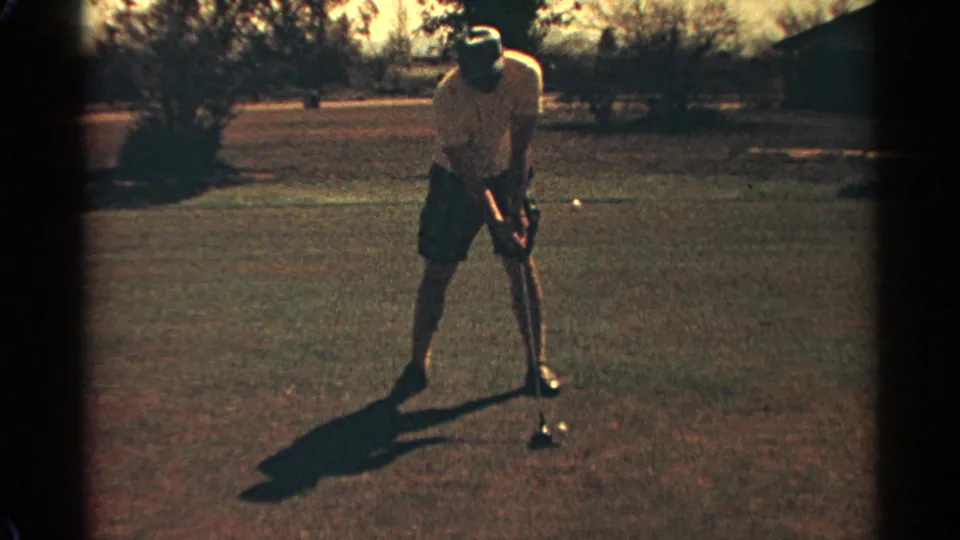 A man in light shorts bends over a golf ball on the Scottsdale course, preparing a shot in grainy 1968 8mm footage.