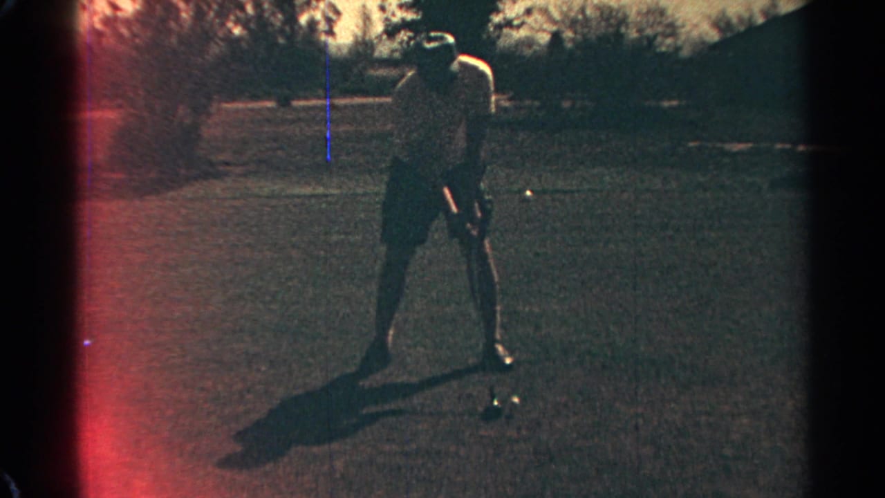 Man addresses a golf ball on grass with a long shadow beside him.