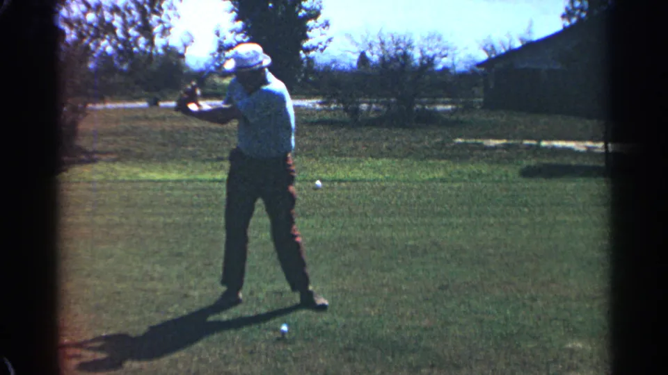 The golfer continues the swing in the 1968 Scottsdale home-movie footage, standing on grass with trees and a low building behind.