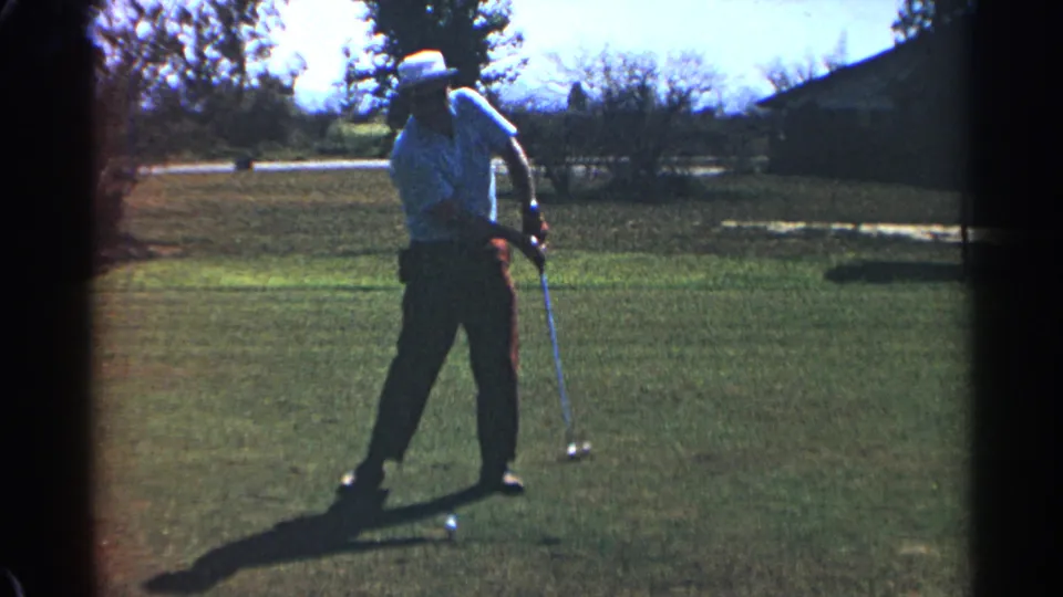 An adult golfer in a white hat stands after the swing with the club lowered, filmed on a sunny Scottsdale golf course in 1968.