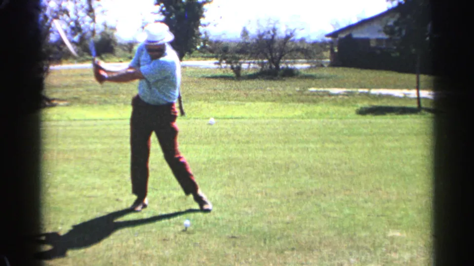 In 1968 Scottsdale, an adult golfer in a white hat, blue shirt, and red trousers starts a drive on a bright grassy course.