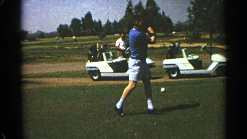 A man on a 1968 Scottsdale golf course finishes his swing near a visible ball, with carts and trees across the fairway.