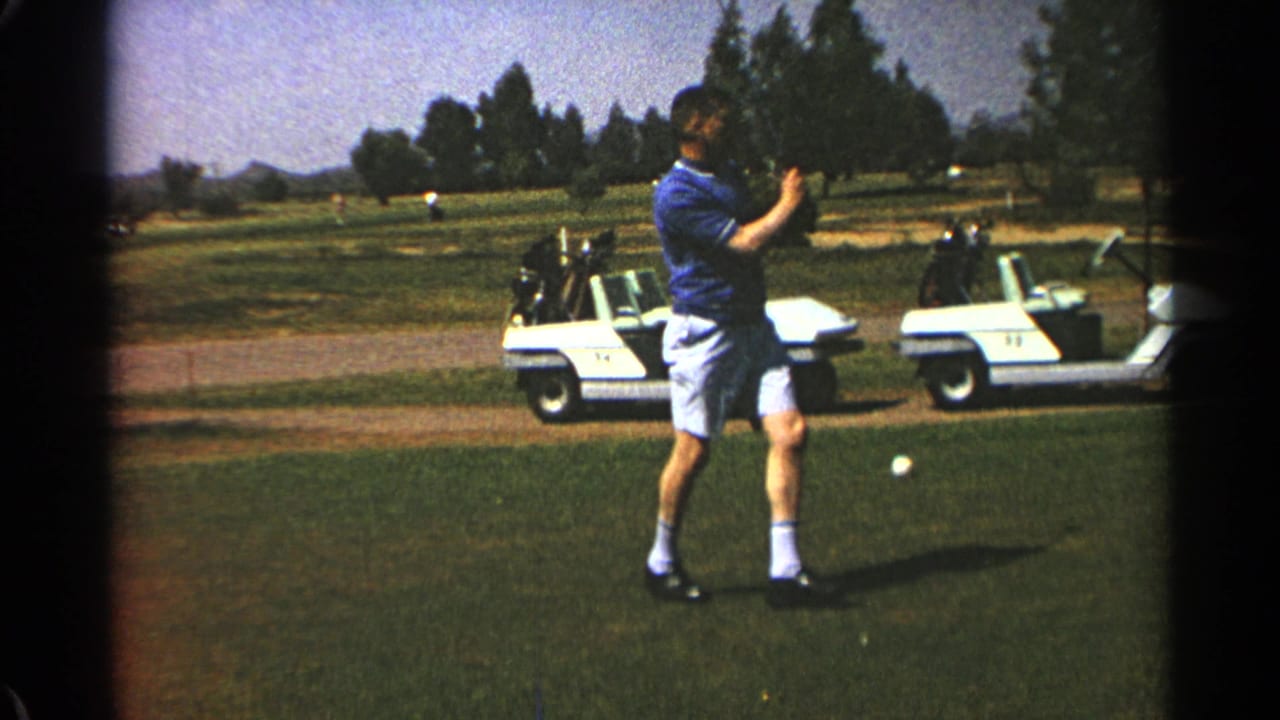 Man in blue shirt swings a golf club on a grass course with white golf carts behind him.