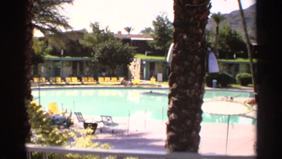 1968 Scottsdale poolside home-movie frame with palm trunks, a turquoise pool, yellow lounge chairs, and low resort-style units.