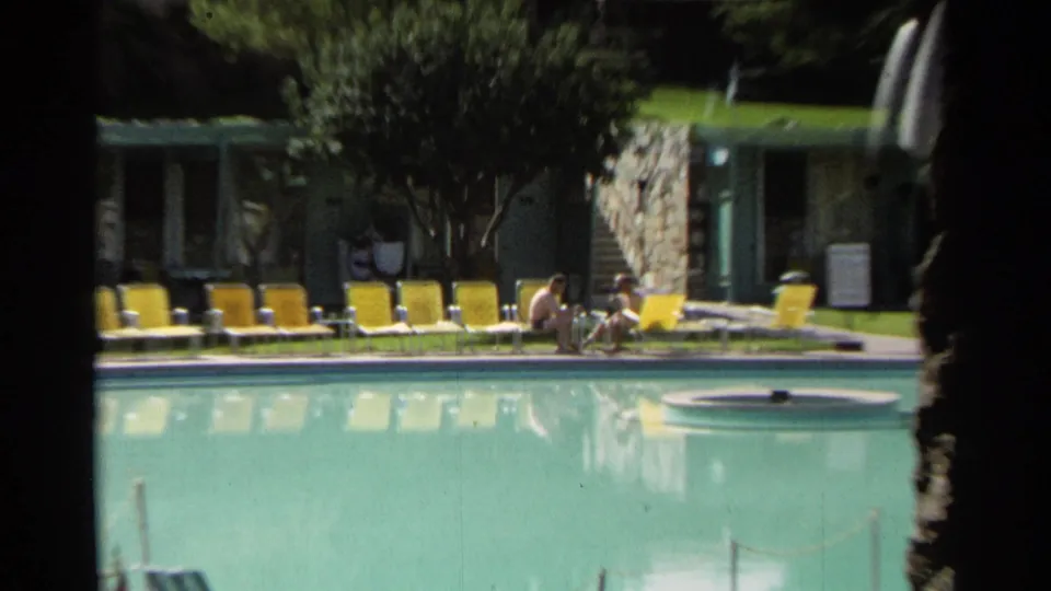 Closer 1968 Scottsdale poolside frame showing two men seated near the water, with yellow lounge chairs and low units behind them.