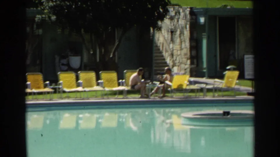 1968 Scottsdale poolside frame with two men seated beside the water, backed by yellow lounge chairs, trees, and low stone-and-glass units.