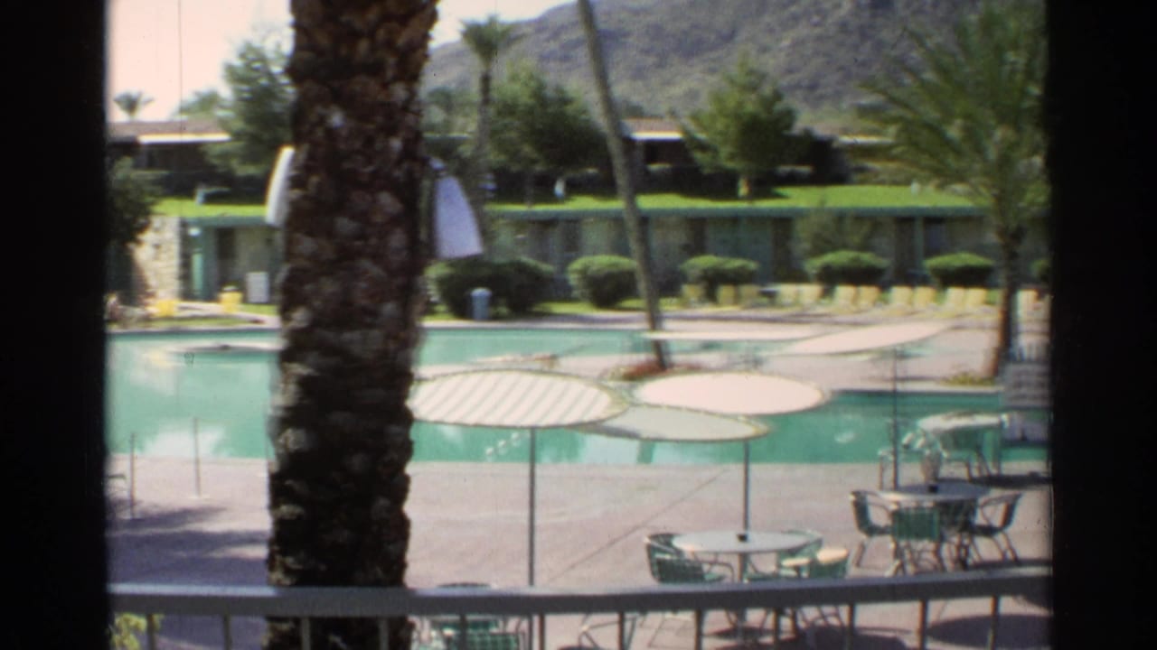 Balcony view of a resort pool with umbrella tables, palm trees, and low buildings beyond.