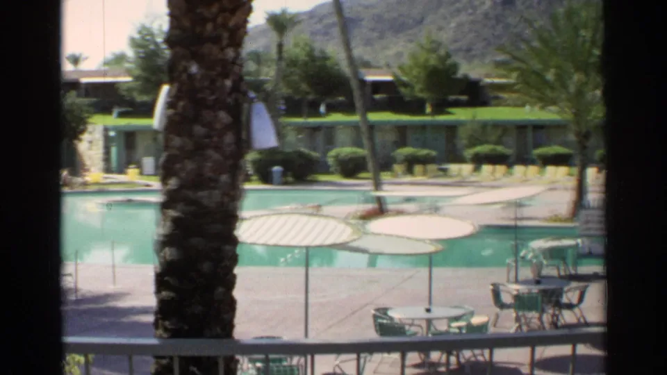 1968 Scottsdale resort or motel pool seen from a balcony, with striped umbrellas, patio seating, palms, and mountains behind low buildings.