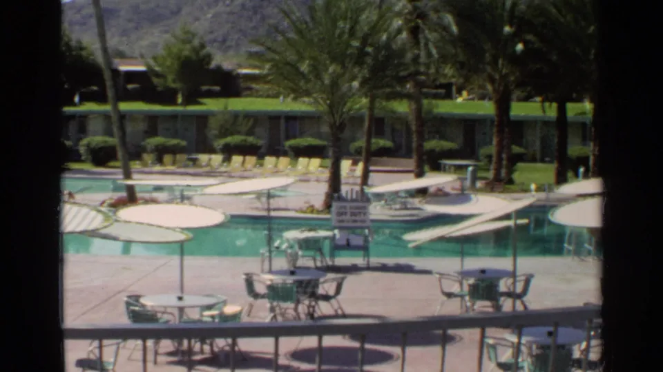 1968 Scottsdale resort pool deck with yellow loungers, umbrella tables, palms, a posted pool sign, and a low building across the water.