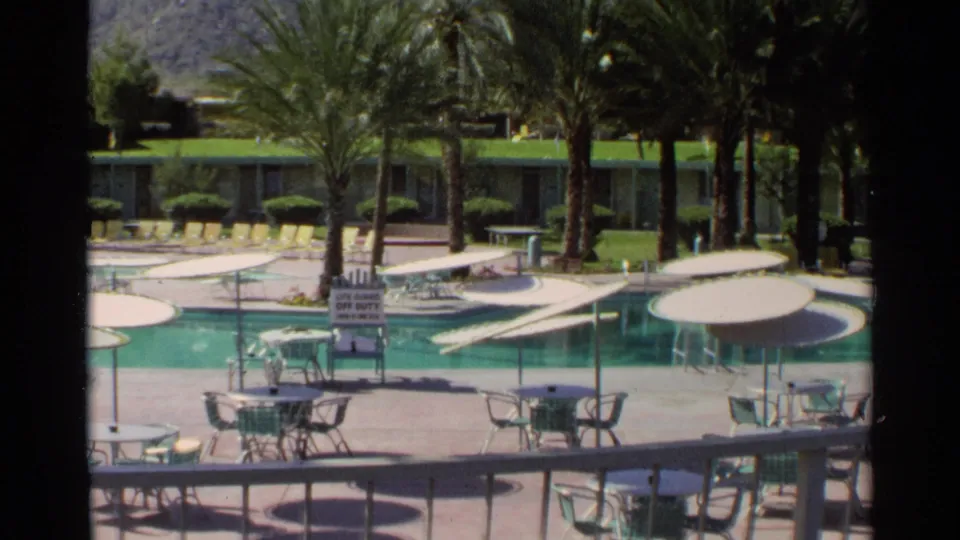 Late-1960s Scottsdale pool scene with palm-lined deck, umbrella tables, patio chairs, yellow loungers, and signage near the water.