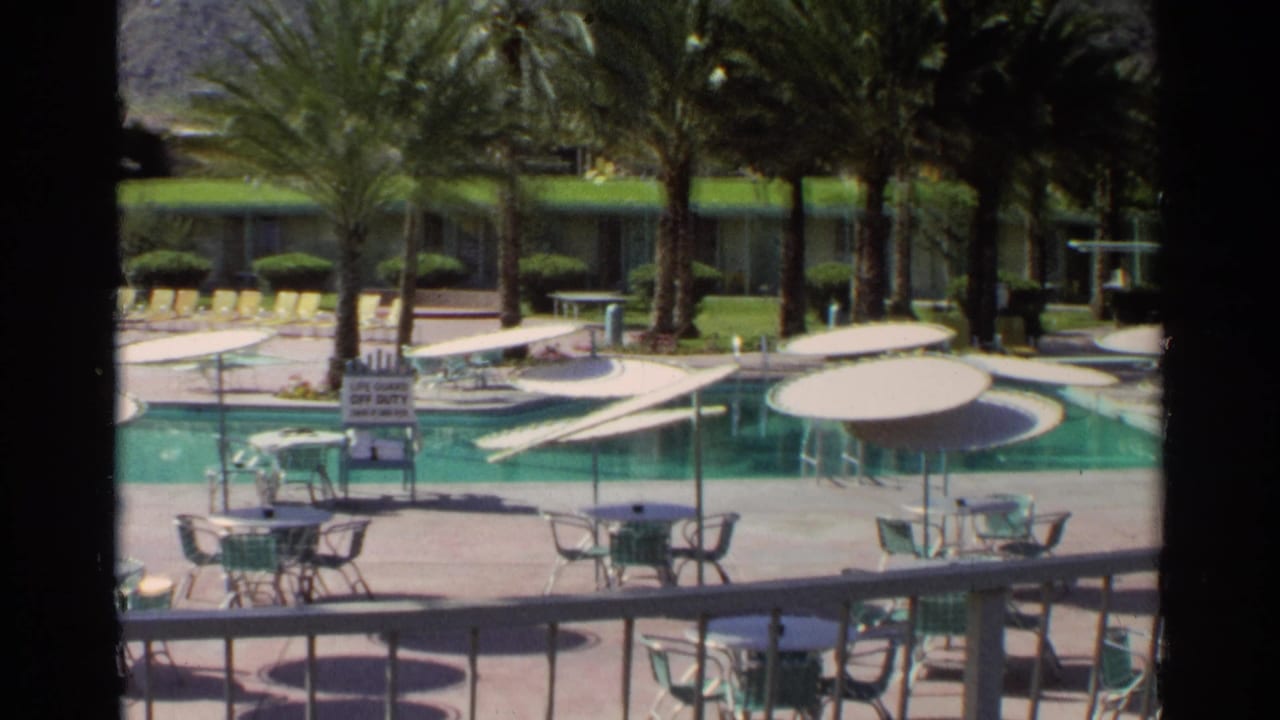 Poolside view with palm trees, umbrellas, tables, chairs, and motel rooms beyond the water.