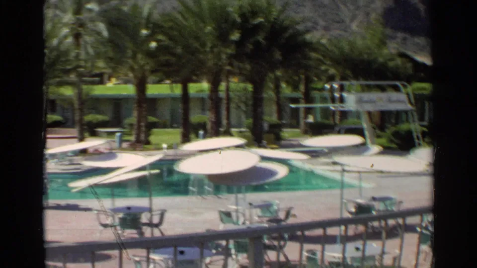1968 Scottsdale poolside frame showing diving boards, white umbrellas, palm trees, patio furniture, and rooms beyond the pool.