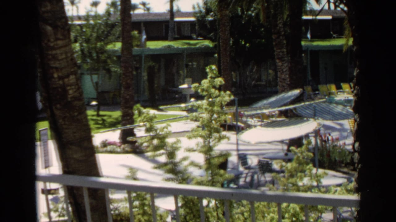 Balcony-framed 1968 Scottsdale poolside scene with palm trunks, white railing, striped umbrellas, motel units, and distant guests.