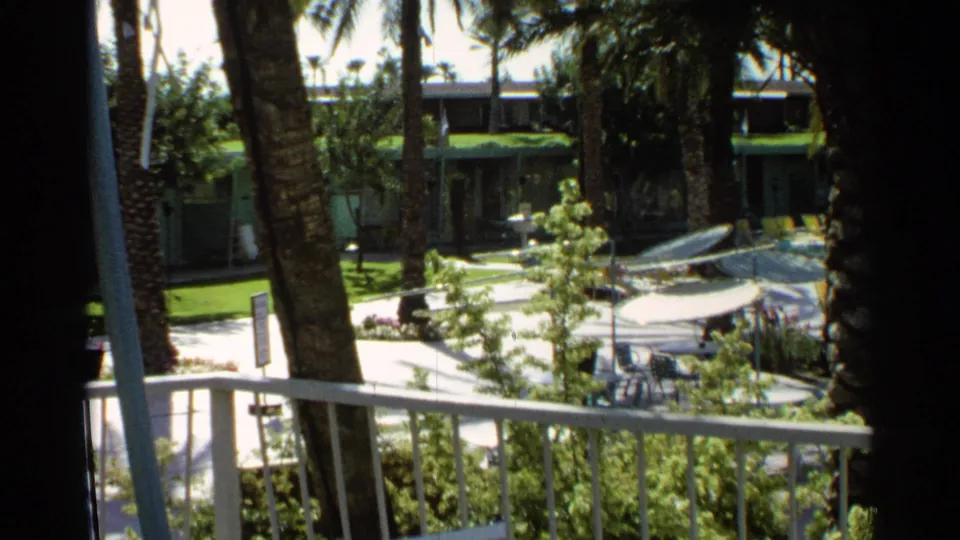 1968 Scottsdale resort courtyard viewed from above, showing a railing, palms, umbrellas, lounge furniture, greenery, and distant pool guests.