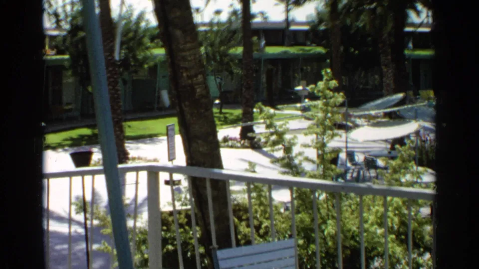 Final 1968 Scottsdale poolside view from a balcony, with white railings, palm trunks, striped umbrellas, lounge chairs, and motel units.